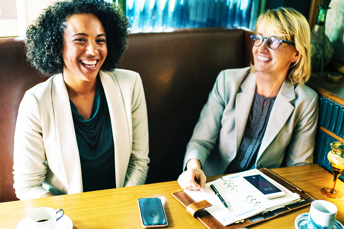 Two smiling business women in suits sit at a table with their phones and diaries.