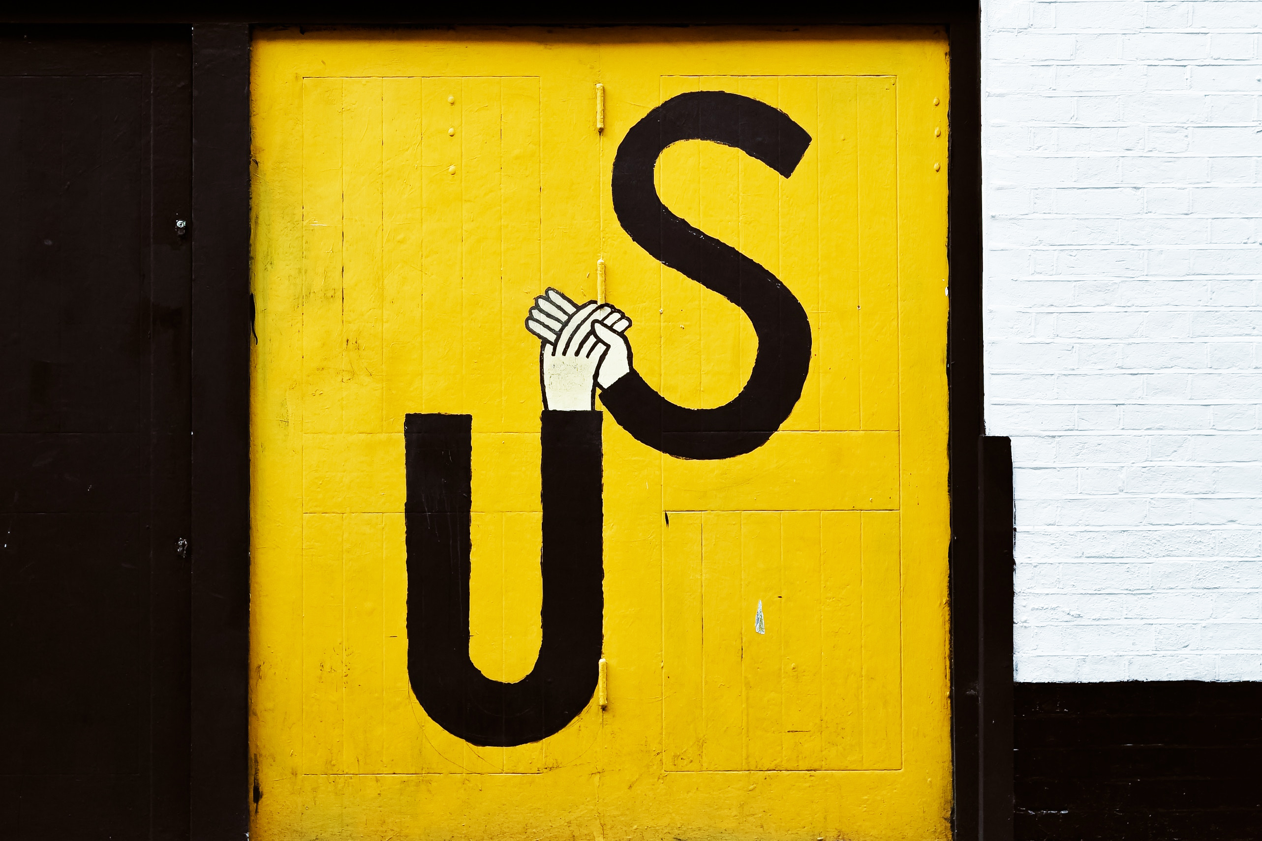A painted street sign that says 'Us' and is designed so that the letter 'u' and the letter 's' are diagonally aligned and joined with two hands