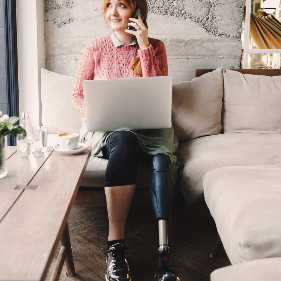 A woman sitting on a couch simultaneously using her mobile phone and laptop, gazing off into the distance.